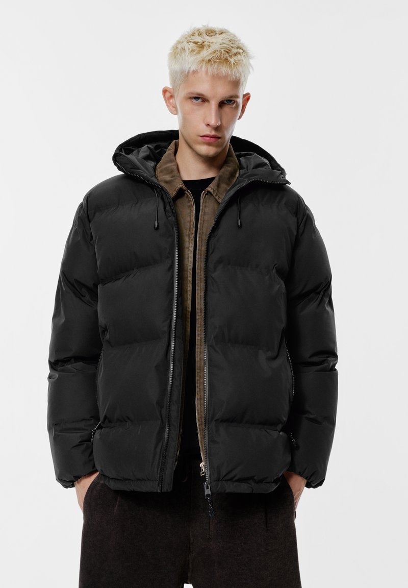 Črna puhasta jakna s kapuco, s prešitimi predeli, sprednjo zadrgo in potegalko. Nošena preko rjave jopice z zadrgo.