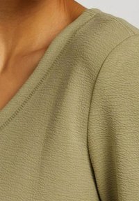 Tessuto di un verde oliva con texture e design a V, che mostra una superficie morbida e dettagli di cucitura visibili lungo il collo e la spalla.