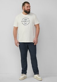 Light cream t-shirt with navy printed text, paired with dark blue pants and beige sneakers. Casual, relaxed fit with short sleeves.
