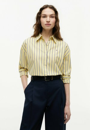 Kobieta o długich falowanych włosach, ubrana w żółtą koszulę w paski zapinaną na guziki, wpuszczoną w granatowe, wysokie spodnie z czarnym paskiem, z rękoma w kieszeniach.