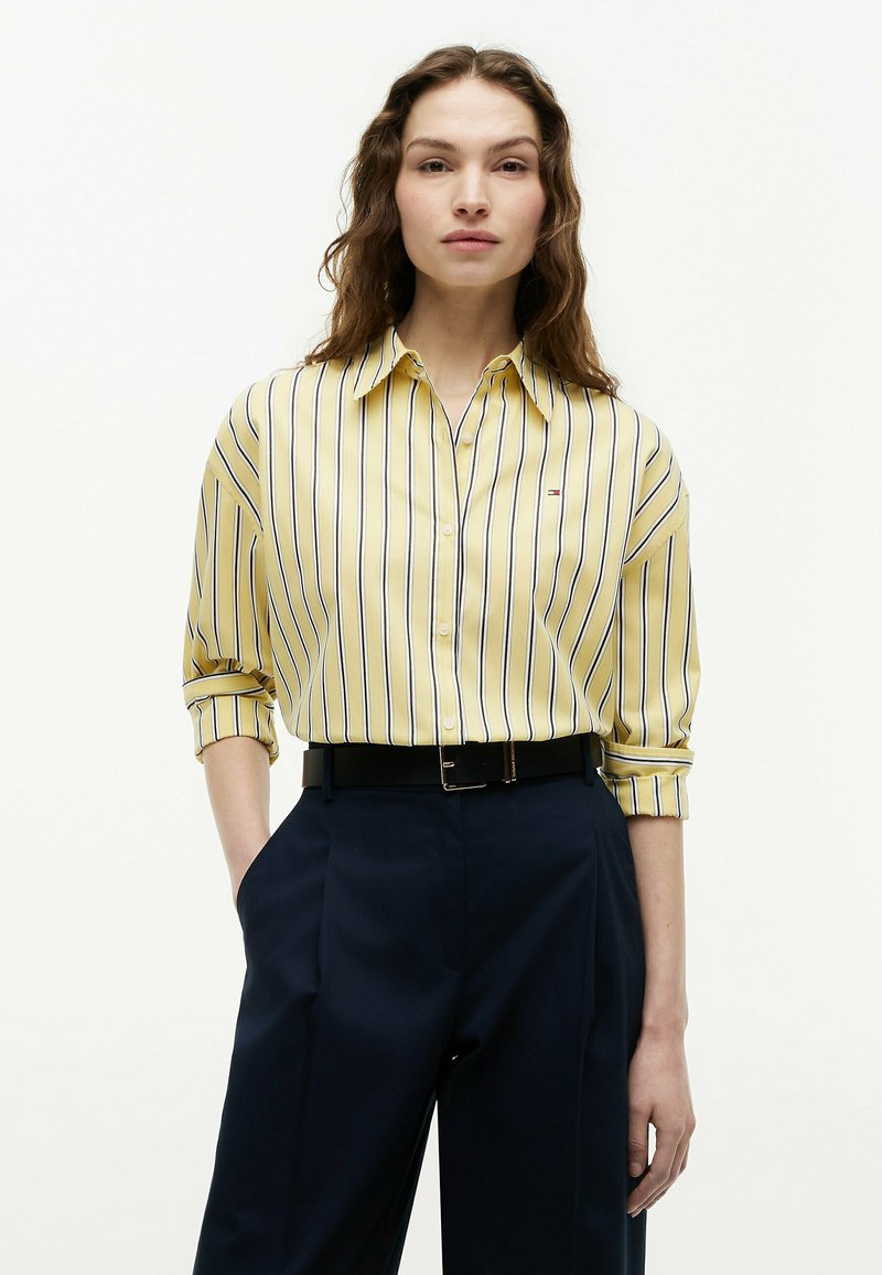 Femme aux longs cheveux ondulés portant une chemise jaune rayée boutonnée, rentrée dans un pantalon taille haute bleu marine avec une ceinture noire, les mains dans les poches.