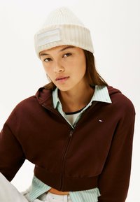 Hoodie corti marrone con zip, con polsini a coste. Indossato sopra una camicia a righe verde e bianca e abbinato a un berretto di maglia beige.