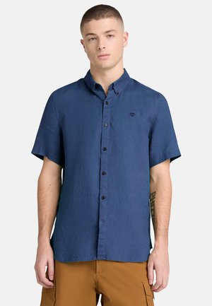 Junger Mann mit kurzen Haaren, der ein navyblaues, kurzärmeliges Hemd mit Knöpfen und braune Cargohosen trägt und vor einem schlichten Hintergrund steht.