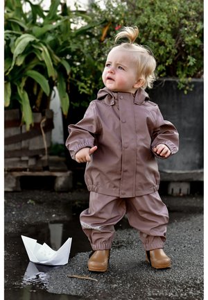 Impermeabile marrone in gomma con pantaloni abbinati, polsini elastici e bottoni. Bambino in piedi in una pozza d'acqua accanto a una barchetta di carta bianca.