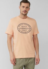Perzikkleurig katoenen T-shirt met korte mouwen, voorzien van een zwart ovale grafische tekstontwerp. Casual pasvorm, aangevuld met gedempte groene broeken.