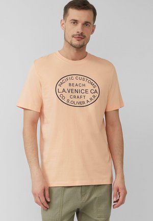 Pfirsichfarbene Baumwoll-T-Shirt mit kurzen Ärmeln, das ein schwarzes ovales grafisches Textdesign aufweist. Lässige Passform, ergänzt durch gedämpfte grüne Hosen.