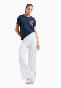 T-shirt di cotone blu navy con maniche arrotolate e grafica circolare; pantaloni bianchi a gamba larga con costine; sneakers bianche. Outfit semplice e casual.