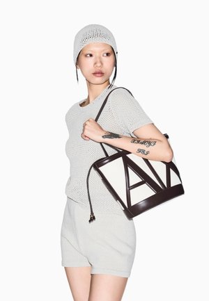 Joven con camiseta de punto clara, pantalones cortos y gorro de crochet a juego, llevando un bolso tote geométrico en blanco y negro colgado del hombro.