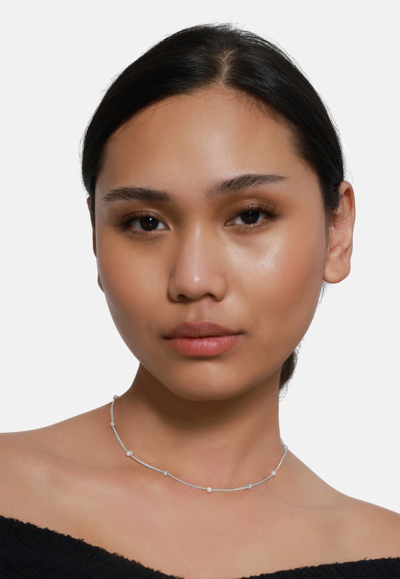Collana d'argento con delicate perline e piccoli dettagli distribuiti uniformemente. Indossata attorno al collo, su una semplice maglia nera e uno sfondo chiaro.