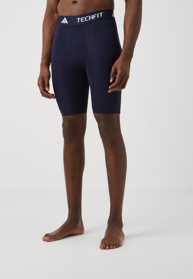 Shorts de compression bleu marine en tissu extensible, avec un logo Adidas Techfit blanc sur la ceinture et une coupe ajustée.