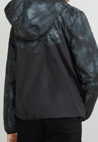 Chaqueta con capucha negra y gris, de tejido texturizado, con un patrón de tie-dye en la parte superior de la espalda y una sección inferior suave, diseño ligero.