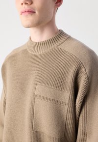 Beige knitted sweater with a high ribbed neckline, textured body, and a patch pocket on the front. Simple, clean design with subtle patterns.