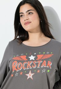 Szara koszula z długim rękawem z grafiką "ROCKSTAR" w kolorach pomarańczowym, białym i neonowym zielonym, z nadrukami gwiazd i skrzydeł z przodu.