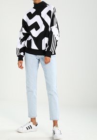 Schwarz-weißes übergroßes Sweatshirt mit kräftigem grafischem Muster, hohem Kragen und gestreiften Ärmeln, kombiniert mit hellblauen Jeans und weißen Sneakern.