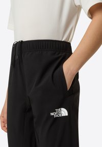 Čierne nohavice s elastickým pásom, s bočným vreckom a bielym logom The North Face na ľavom stehne. Hladká textúra.