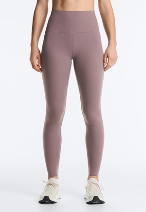 Nő, aki mályvaszínű, magas derekú leggingset és fehér tornacipőt visel, karjai lazán az oldalán nyugszanak egy egyszínű háttér előtt.