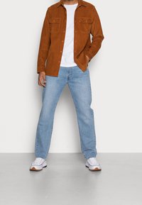 Camisa de pana marrón con dos bolsillos en el pecho, usada sobre una camiseta blanca, combinada con vaqueros azul claro y zapatillas blancas.