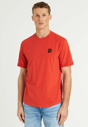 T-shirt rossa in cotone con maniche corte, colletto rotondo e un'etichetta con logo nera sul petto. Indossata con jeans azzurri chiari.