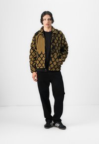 Fuzzy olive-green jacket with a black geometric pattern, paired with a black T-shirt and black pants, worn with black sneakers.