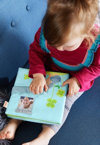 Soft fabric book in light blue, featuring colorful illustrations, a plastic photo pocket, and a tactile closure. Child’s hand interacts with the book.
