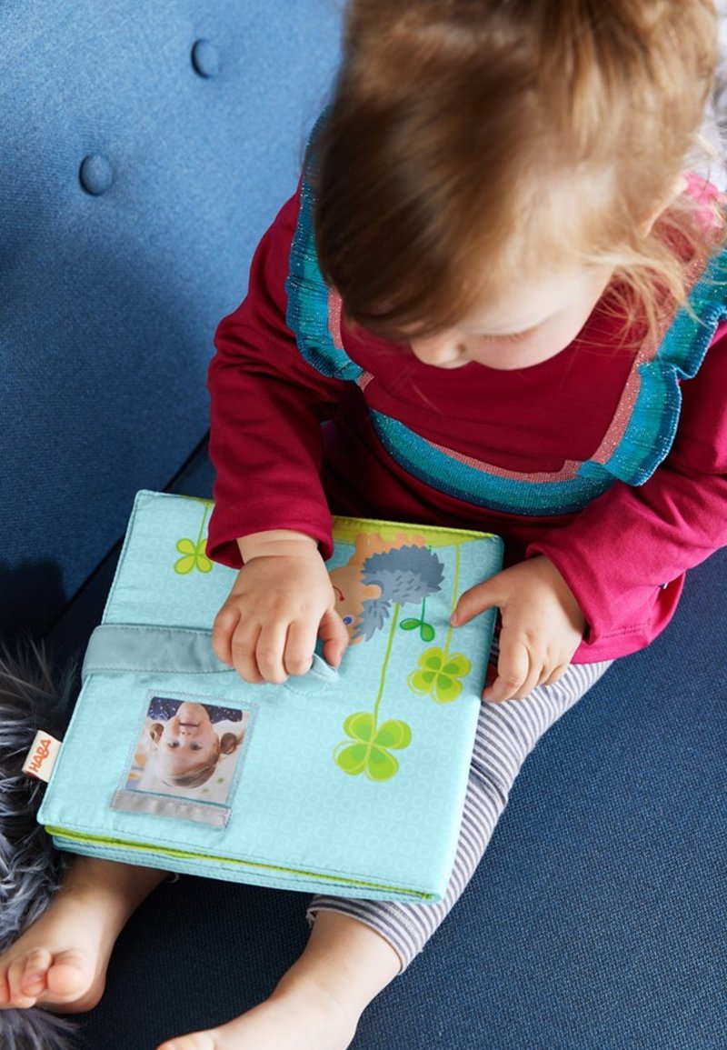 Soft fabric book in light blue, featuring colorful illustrations, a plastic photo pocket, and a tactile closure. Child’s hand interacts with the book.