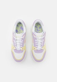 Sneakers Lacoste mają białą skórzaną cholewkę z akcentami z zamszu w kolorze lawendy, różowym i żółtym, teksturowane sznurówki oraz wkładkę w kamuflażowy wzór.