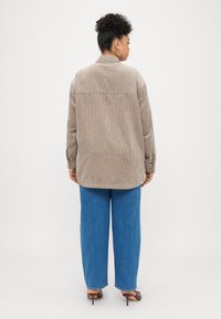 Rudas korduroy marškininė striukė su užsegamu apykakle, ilgomis rankovėmis ir išlenktu kraštu, derinama su mėlynomis džinsinėmis plačiomis kelnėmis.