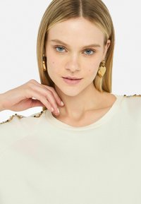 Felpa beige con dettagli decorativi in perline sulle spalle. Il modello ha una carnagione chiara e indossa orecchini a forma di cuore dorati. Mani che sfiorano delicatamente il collo.