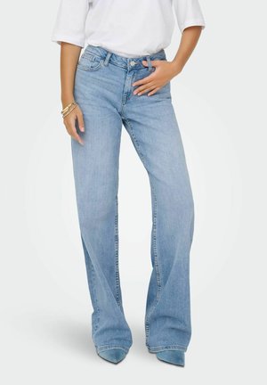 Kvinde iført lyseblå vidde jeans og spidse blå sko, hvid langærmet top, med den ene hånd på linningen og armbånd på håndleddet.