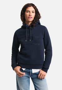 Felpa blu navy realizzata in misto cotone, con cappuccio regolabile, logo in rilievo e polsini e orlo a coste. Indossata con jeans chiari.