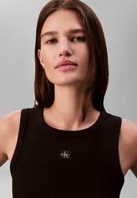 Jonge vrouw met steil bruin haar, gekleed in een zwart mouwloos topje met een klein Calvin Klein-logo op de borst, neutrale grijze achtergrond.