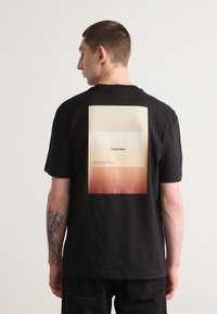 Musta puuvillapaita, jossa on suuri graafinen kuvio selässä, joka vaihtelee kermanväristä oranssiin ja sisältää tekstin "Calvin Klein."