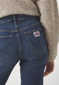 Mörkblå denimjeans med hög midja, två bakfickor med en vit etikett, och distinkt sömnad som bildar en V-form. En texturerad tröja är delvis synlig.