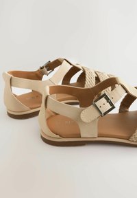 Beige sandals with woven straps, leather material, adjustable buckle closure, flat sole, and smooth interior. Suitable for casual wear.