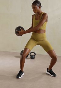 Atletska odjeća u maslinasto zelenoj boji, koja se sastoji od skraćenog tankog topa i kratkih hlača visokog struka. Crna lopta za vježbanje drži se obema rukama, sportske cipele.