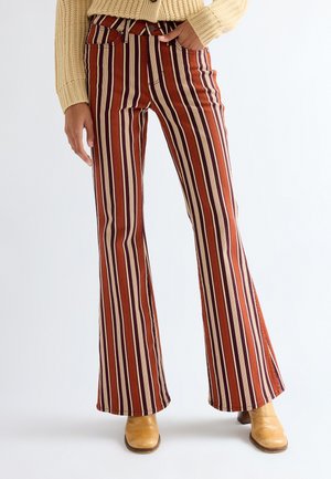 Persona che indossa pantaloni a vita alta e a zampa con righe verticali rosse, marroni, crema e viola e stivaletti color cuoio, in piedi su sfondo bianco.