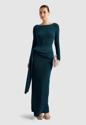 Türkisfarbenes, funkelndes Langarmkleid mit figurbetontem Schnitt, seitlichem Knotendetail und bodenlangem Saum. Kombiniert mit einer silbernen Clutch und minimalistischen Sandalen.