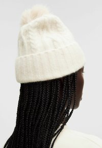 Valkoinen neulottu pipo, jossa on ribbikuvio ja suloinen pompomi ylhäällä. Suunnittelussa on käytetty cable-knit -kuviota.