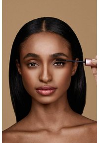 En svart mascaraborste appliceras på fransarna, hålls av en hand. Modellen har slät hy, rakt svart hår och en neutral bakgrund.