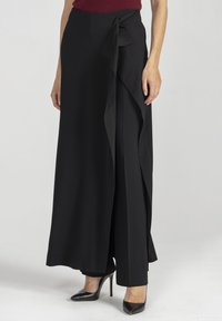 Długa czarna spódnica z gładkiej tkaniny, z przodu z drapowaniem, wiązana w talii, o zwiewnej sylwetce. Połączona z czarnymi, spiczastymi szpilkami.