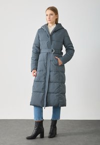 Γυναίκα που στέκεται φορώντας ένα μακρύ γκρι επενδυμένο παλτό με ζώνη και κουκούλα, ανοιχτούς μπλε τζιν και μαύρες μποτές με τακούνι, σε ουδέτερο φόντο.