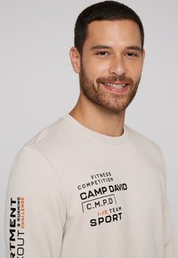Lys beige sweatshirt med rund hals, prydet med sort og orange teksttryk. Materialet ser glat ud med en afslappet pasform og ribbede manchetter.
