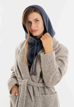 Grijze, textuurrijke jurk met een ceintuur en zakken, gedragen met een blauwe gebreide capuchon met trekkoorden en houten accenten.