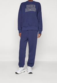 Sudadera de algodón azul marino con el logo de "UNDER ARMOUR", combinada con pantalones jogger a juego. Cuenta con puños y cintura de canalé. Zapatillas blancas.