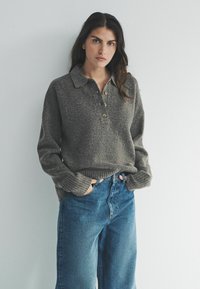 Šedý pletený sveter s golierom, troma gombíkmi a rebrovanými manžetami, skombinovaný s vysokými modrými džínsami. Jednoduchý a neformálny dizajn.