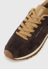 Pruun nahkainen tennisjalanik, millel on beežid nahkaaktsendid, beežid lipsud, ümar varvas ja tekstureeritud kummitald. Michael Korsi bränding on nähtav.