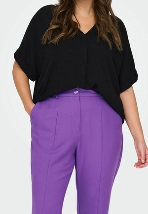 Vrouw draagt een loszittende zwarte top met korte mouwen en hooggetailleerde paarse op maat gemaakte broek met handen in de zakken tegen een witte achtergrond.