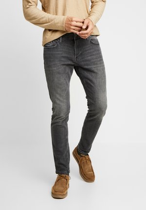 Person iført mørkegrå slim-fit jeans, tan langærmet skjorte og brune ruskinds snøresko står med hænderne foldet.