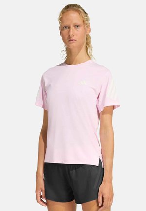 Donna con capelli chiari e ricci che indossa una maglietta Adidas rosa chiaro e pantaloncini sportivi neri, in piedi davanti a uno sfondo semplice.