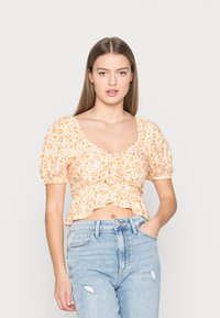 Top cropped floral em branco e laranja, com mangas bufantes, decote quadrado e frente franzida com uma bainha em babados. Usado com jeans claros.